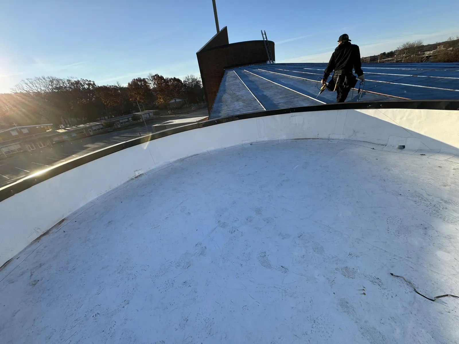 A finished white flat roof with a curved parapet and metal coping, with a blue standing seam roof section and a brick tower behind it