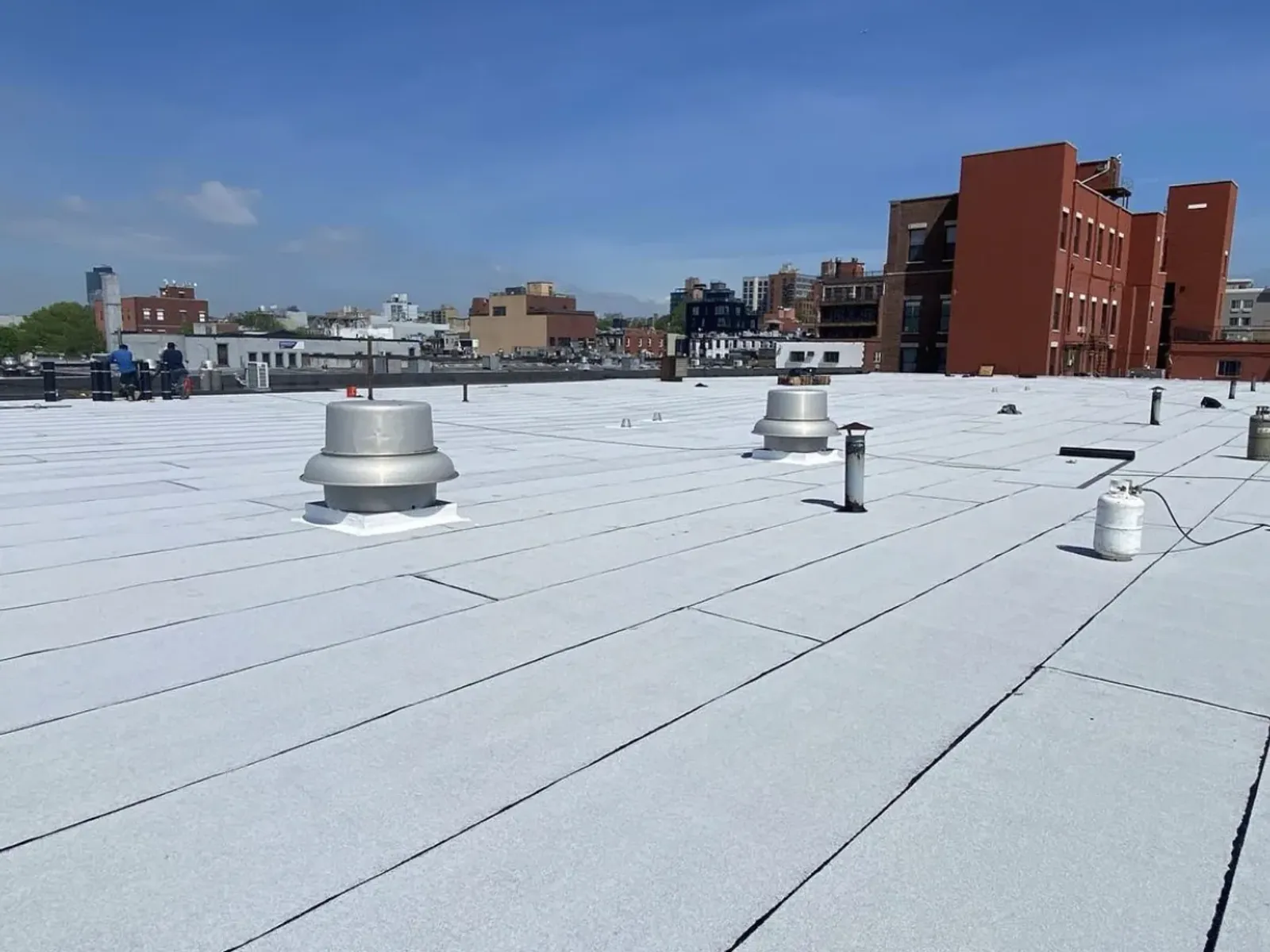 A finished white membrane flat roof with metal vents, with city buildings and a red brick block in the background