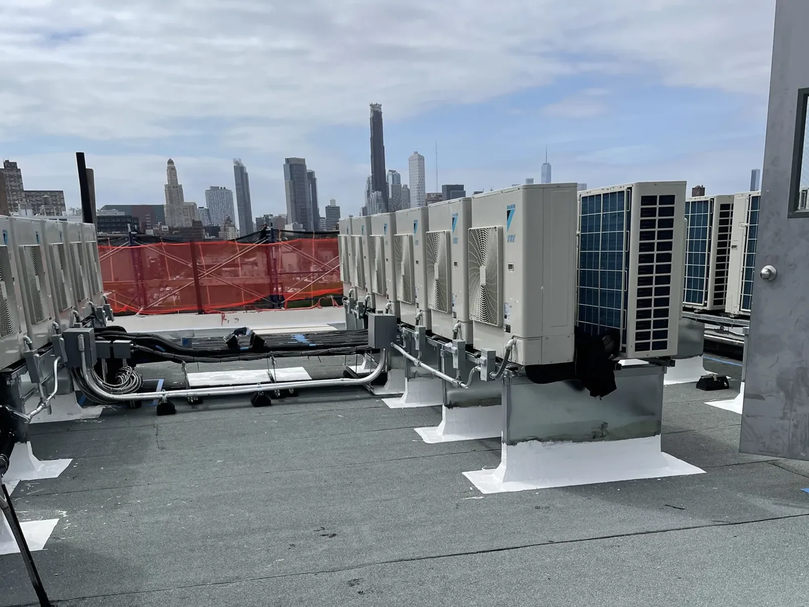 A row of large white rooftop air conditioning units on painted curbs on a finished grey commercial roof, with a city skyline behind