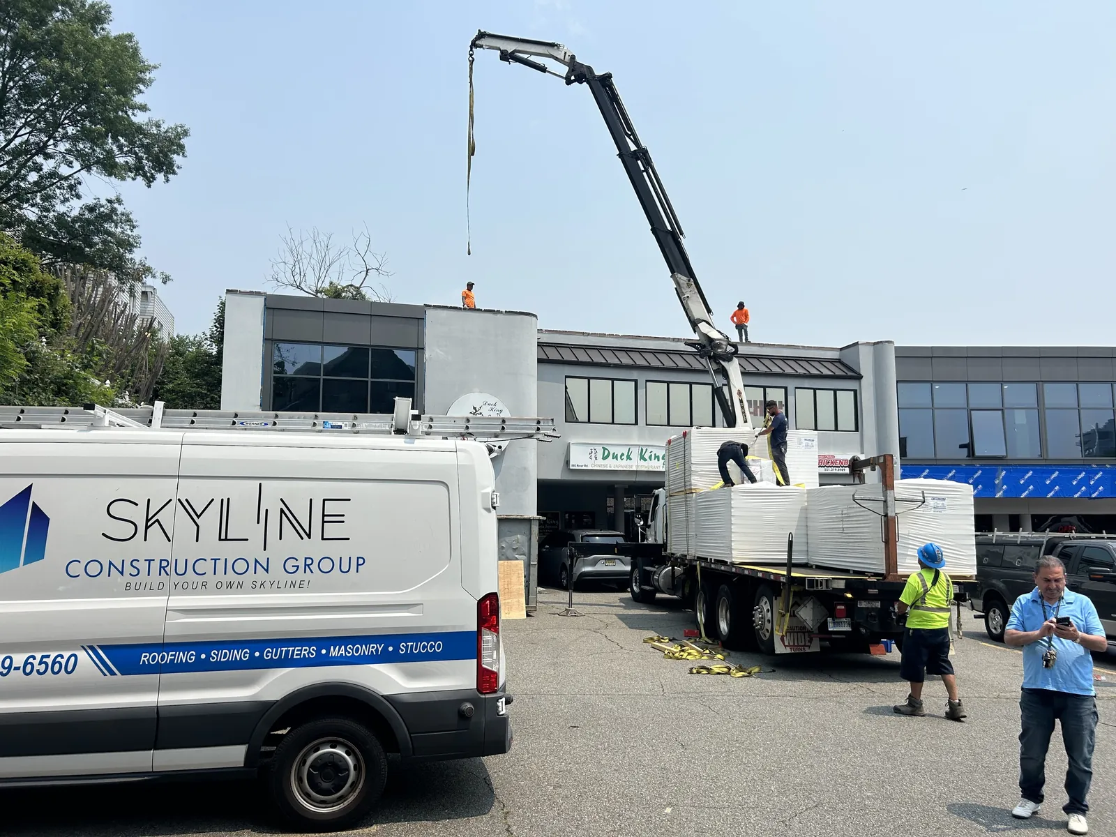 A Skyline Construction Group van in front of a commercial building while a crane lifts panels off a flatbed