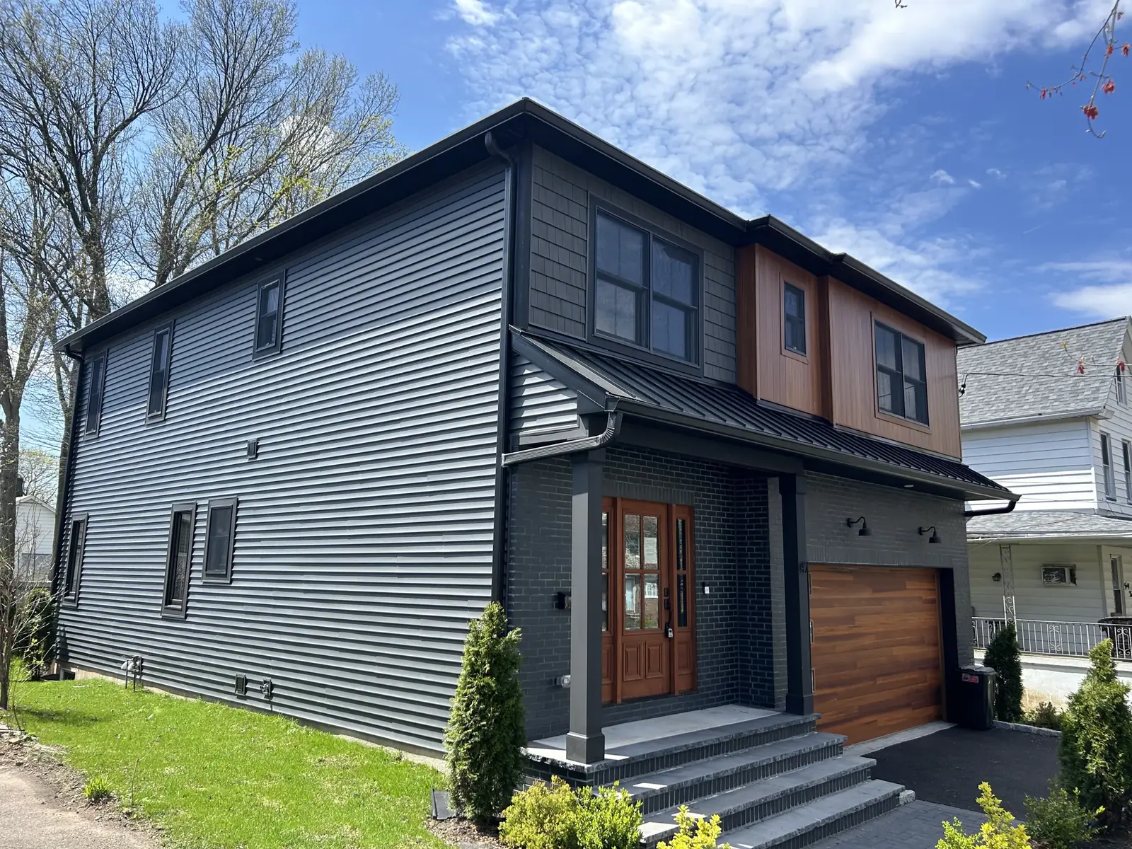 A finished modern home with grey horizontal metal siding, a black standing seam roof and wood tone garage and entry accents