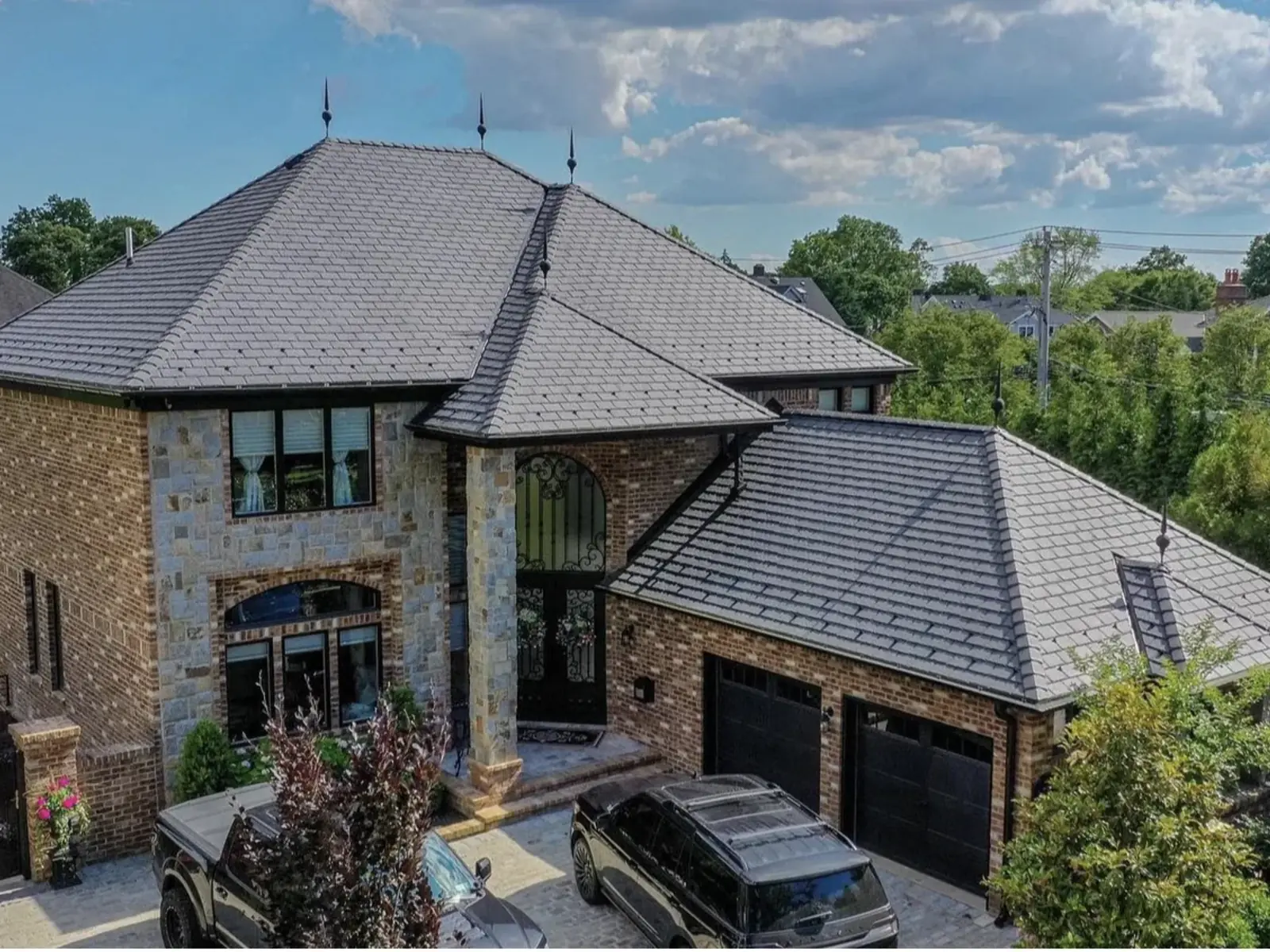 Aerial view of a finished slate look shingle roof on a large stone and brick home with cars in the driveway