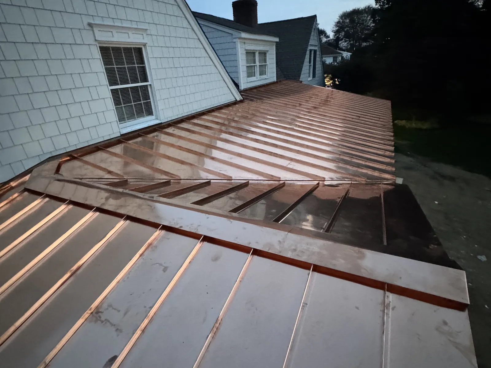 A finished copper standing seam roof panel on a shingle style home at dusk
