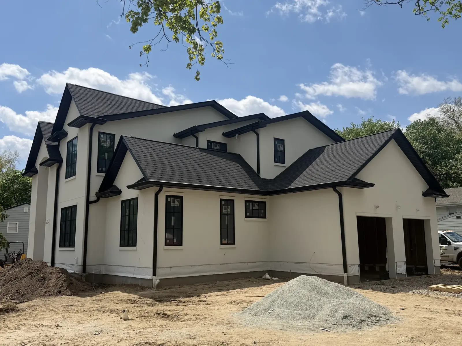 A large white stucco two story home with black trim and gables, finished walls and roofline, with a gravel mound and a truck in the dirt yard